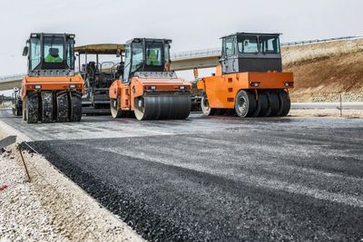Three orange road rollers compacting newly laid asphalt on a highway.