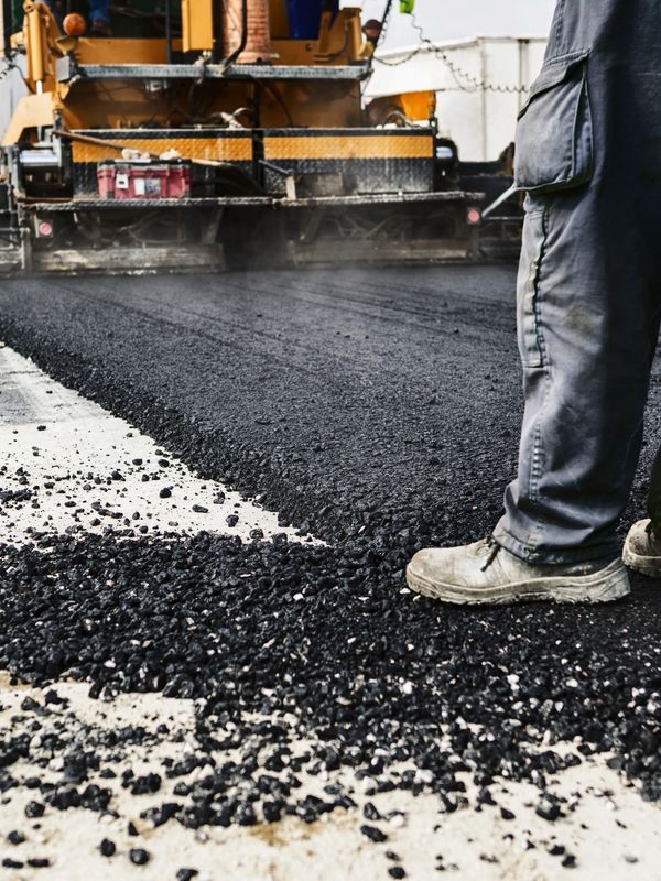 Construction worker standing on freshly laid asphalt with paving machine in background.