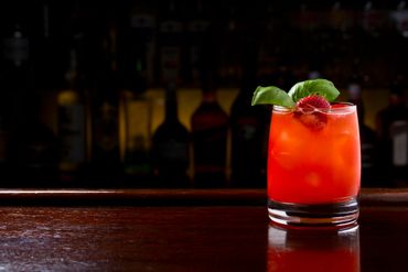 Bright red cocktail garnished with basil and strawberry on a wooden bar.