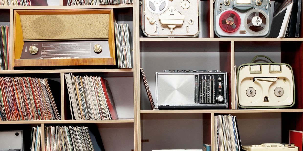 A collection of vintage radios, reel-to-reels, and vinyl records on wooden shelves.