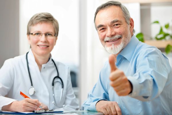Smiling elderly man giving thumbs up with a female doctor in the background.