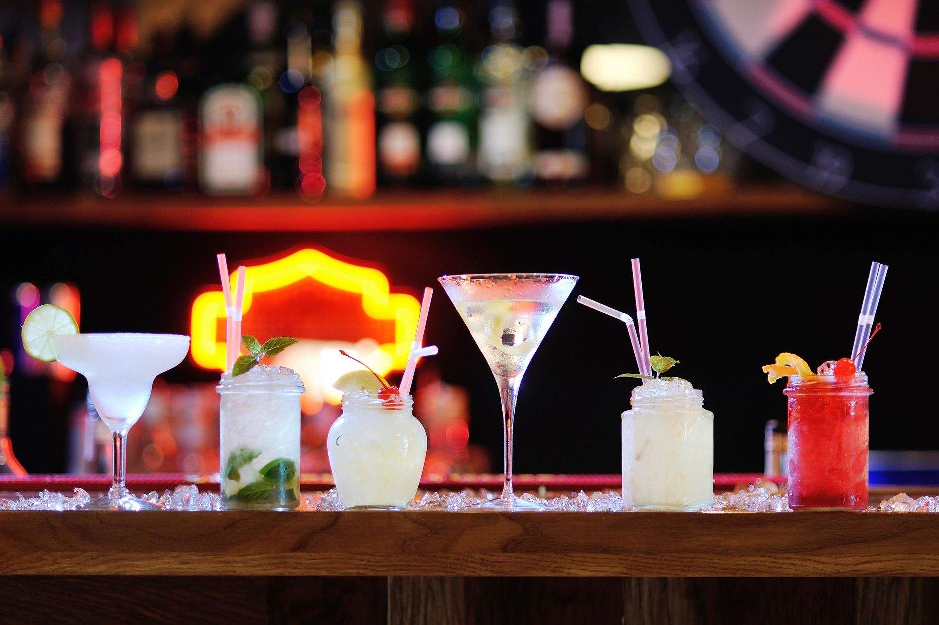 Colorful cocktails lined up on a bar with neon lights in the background.