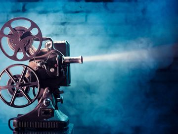 A vintage film projector stands on a pedestal, projecting light through its lens creating a beam that pierces the blue-tinted misty background. The projector has two reels, one at the top and one on the side, with film running through its mechanism.