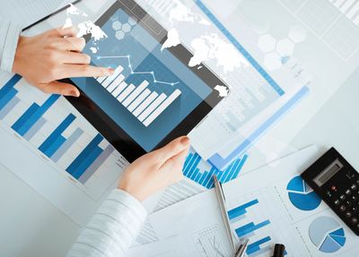 Hands on an iPad depicting graphs, surrounded by papers with charts and a calculator. 