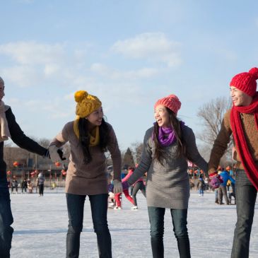 Four friends ice skating outdoors, holding hands and smiling.