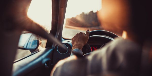 An over-the-shoulder view of a driver with a hand on the steering wheel and wearing a seat belt.