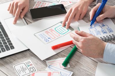 Hands of two people using a computer and markers to draw tables on papers. 