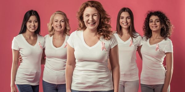 Group of diverse women wearing white shirts with pink ribbons, smiling against a pink background.