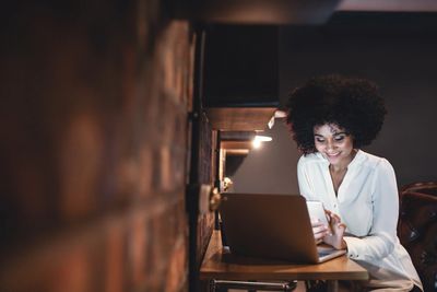 Woman smiling while using phone and laptop in cozy workspace.