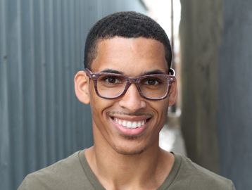 Young man with glasses smiles warmly in an outdoor setting.