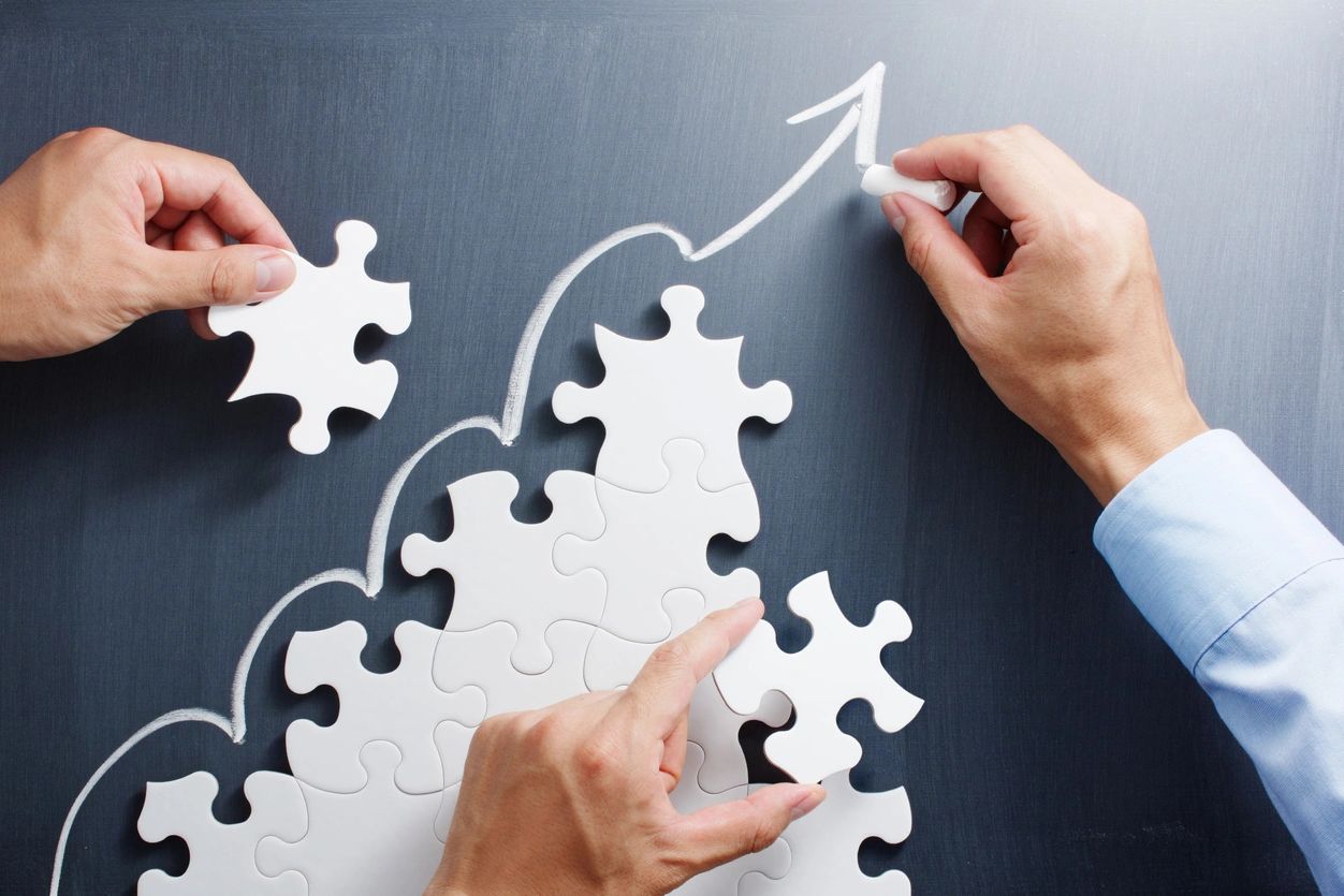 Hands assembling puzzle pieces with upward arrows drawn on a chalkboard.