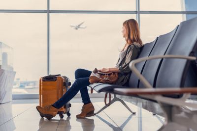 A woman sits waiting at an airport terminal with luggage as a plane flies outside.