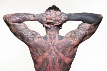 Man showing extensive black and gray tattoos covering his back and arms.
