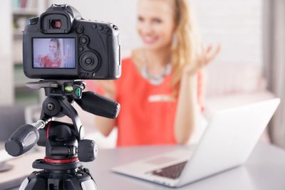 A woman records a video with a DSLR camera while sitting at a laptop.