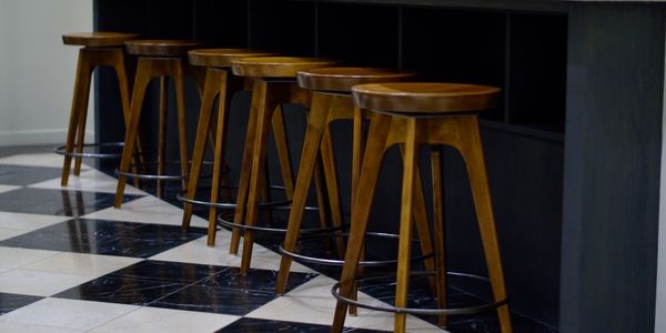 Row of wooden bar stools lined up against a black counter on a checkered floor.
