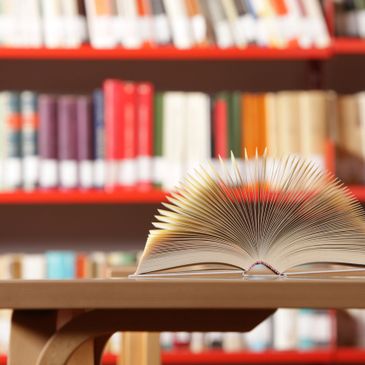 Color photo of wide-open book on a table, with full bookshelves in the background