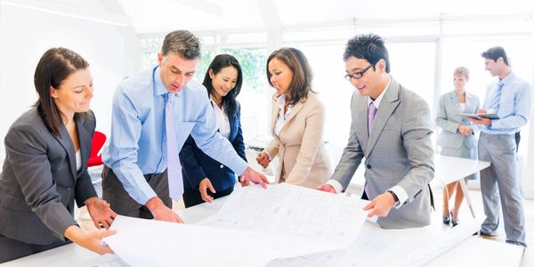 A group of 6 individuals in suits all gathered around building plans (vague) and discussing. 