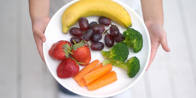 A plate with banana, grapes, strawberries, broccoli, and carrot sticks held by hands.
