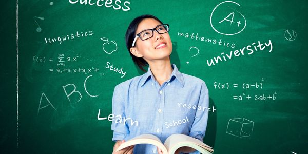 A student holding a book in front of a chalkboard with educational words and formulas.