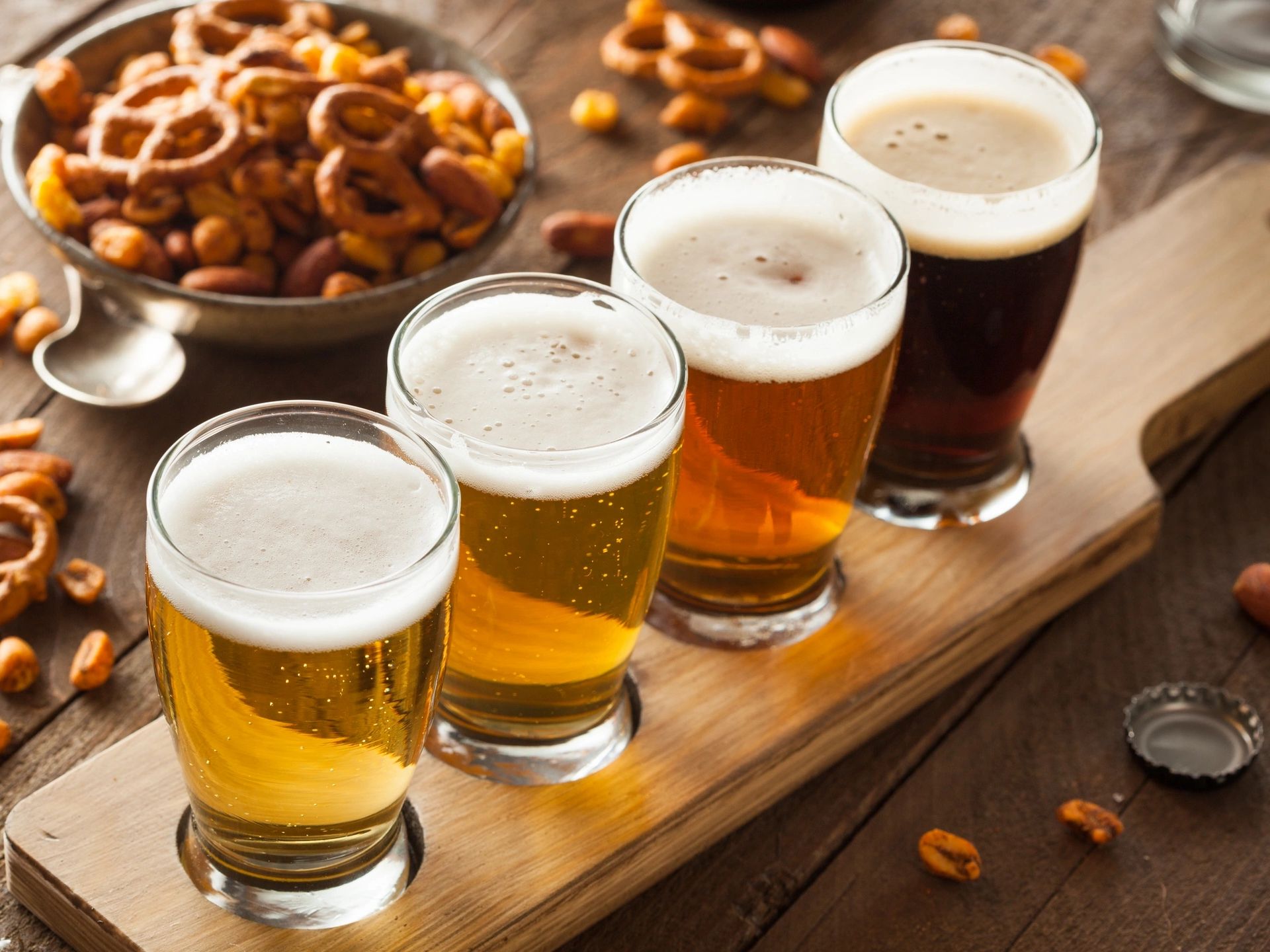 Four beers with foam on a wooden tray and a bowl of snacks.