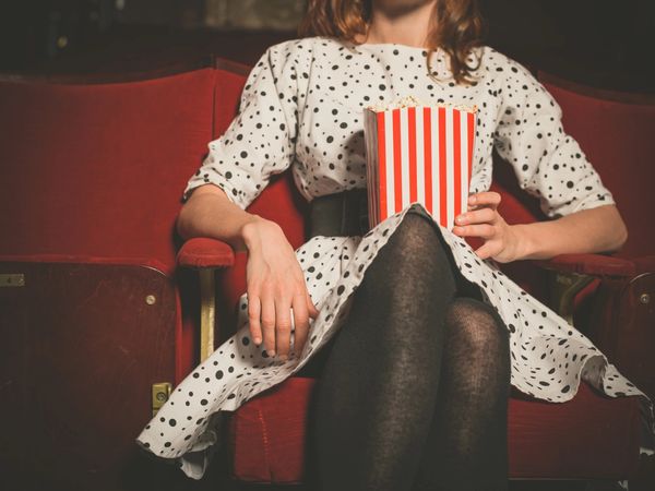 Ung kvinne med hvit polkadot kortermet sekstitalls-kjole som sitter i en kino med popcorn i fanget