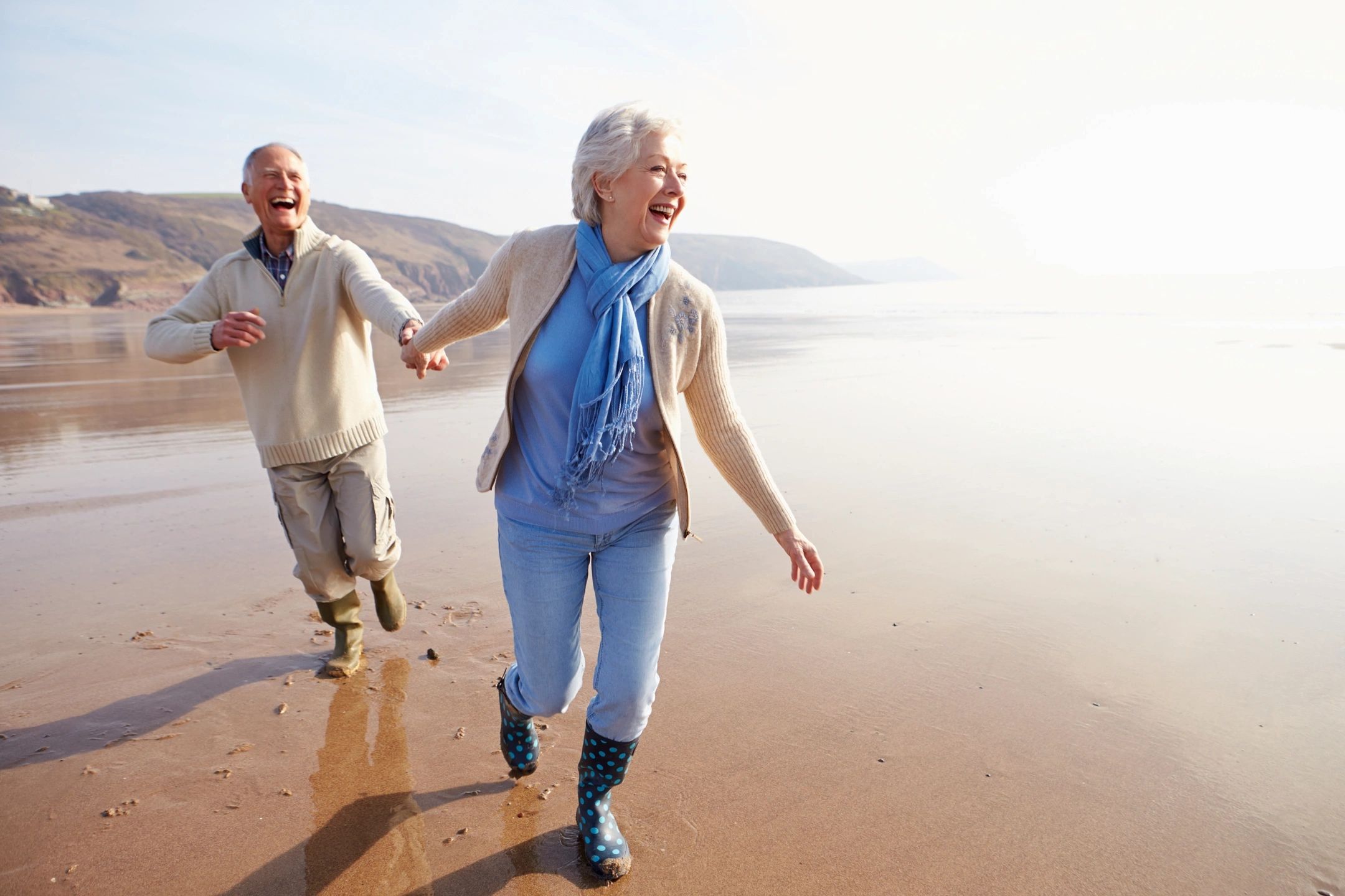 An older couple laughing and holding hands on the beach.