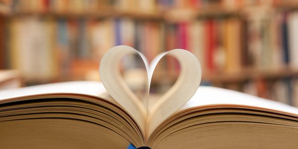 Open book with two pages turned inward to make a heart, blurry library in the background.