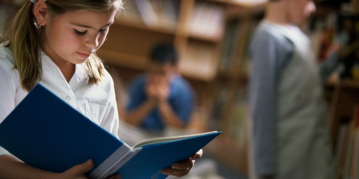 Young girl reading a book in a library with other children in the background.