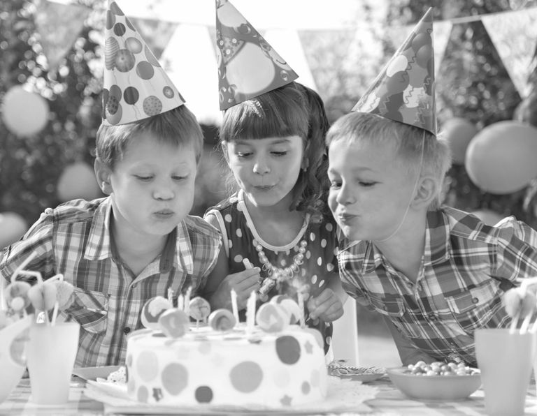 Three children in party hats blowing out the candles on a birthday cake