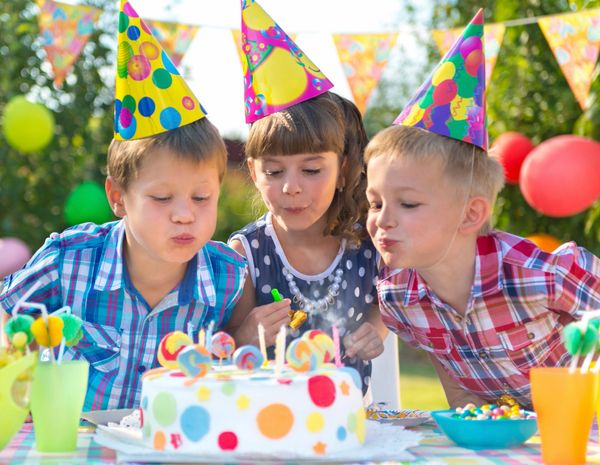 Three children in party hats blowing out birthday cake candles outdoors.