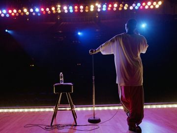 Comedian standing on stage with microphone and stool under bright lights.