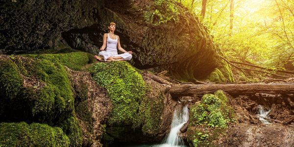 A woman doing yoga on a large rock in the forest in front of a flowing spring