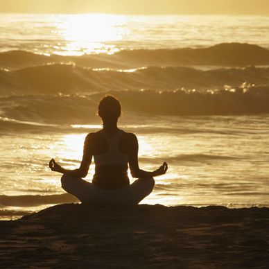 Person meditating in lotus pose by the ocean at sunset.