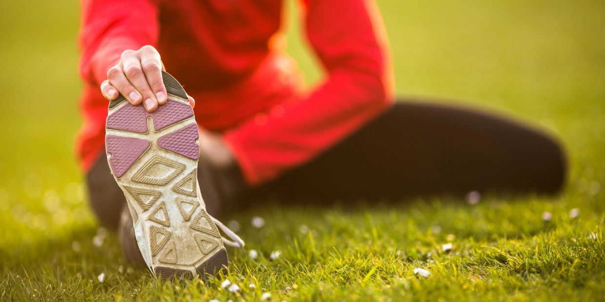 Person stretching on grass, holding foot in workout shoe.