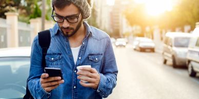 Man in denim jacket using phone and holding coffee on street.