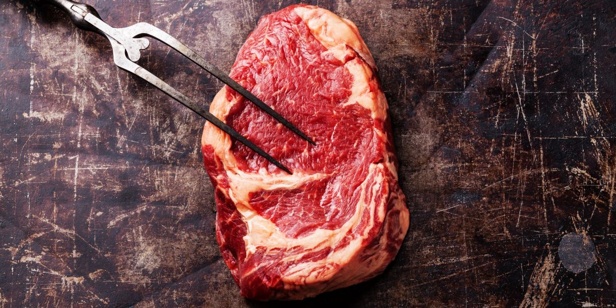 Raw marbled steak held by a meat fork on a rustic surface.