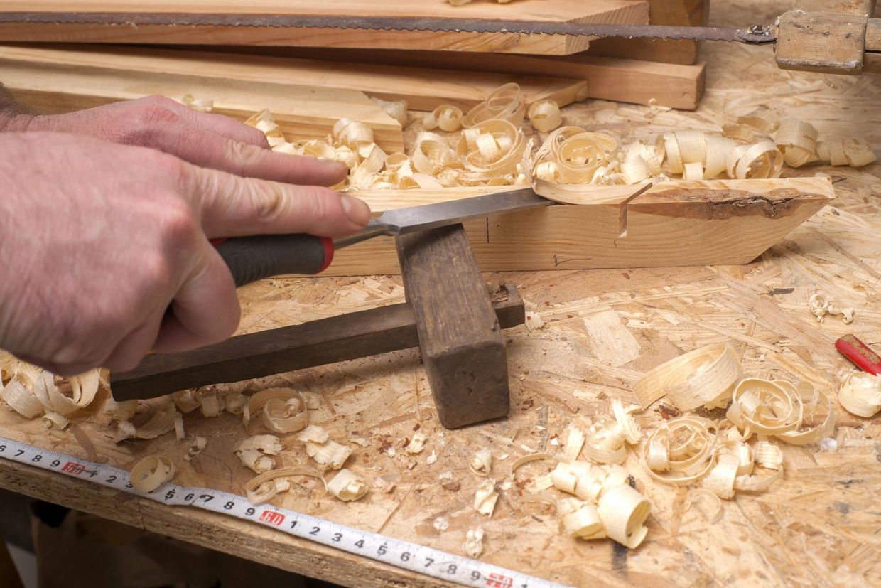 A person carving wood with a chisel and mallet on a cluttered workbench.