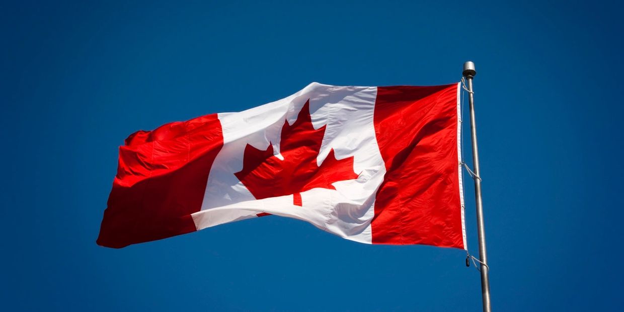 The Canadian flag at the top of a pole that is blowing in in the wind. 