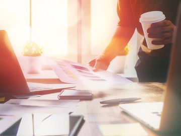 Person analyzing documents with a coffee cup in hand in a sunlit office.