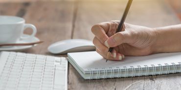 Person writing in a spiral notebook beside a keyboard and coffee cup.