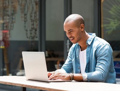 A man smiling while working on a laptop outdoors.