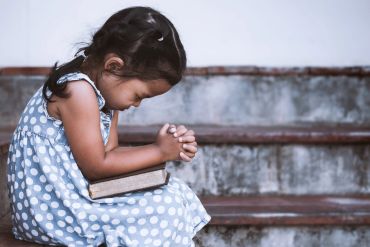 A young girl in a blue polka dot dress praying with a book on her lap.