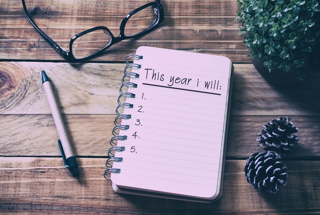 A notepad with "This year I will..." written on it, sit on a tabletop along with a pen and eyeglasses.