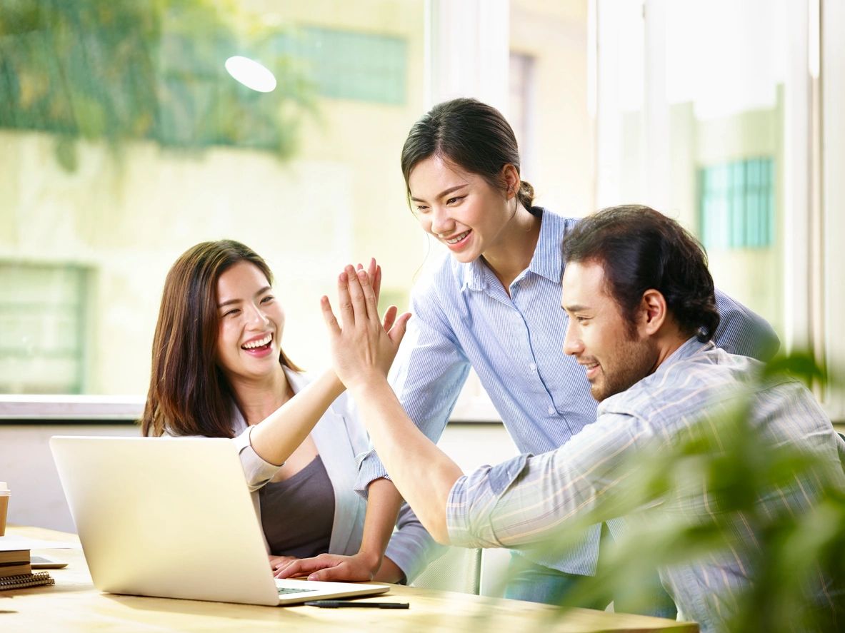 Man and woman do a high five smiling and another woman smiles stands in middle looking at laptop
