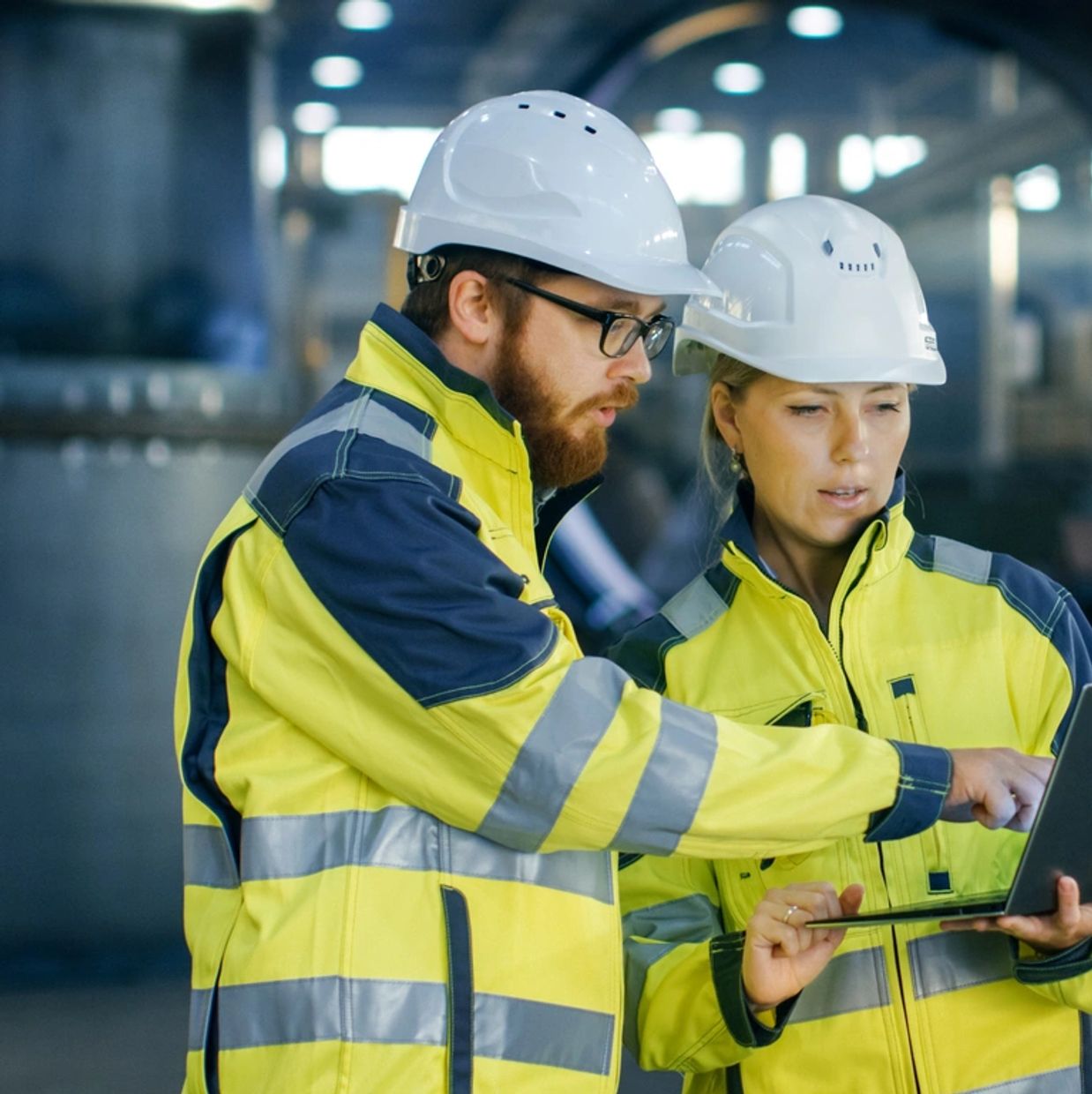 Two engineers in safety gear discussing plans using a laptop in an industrial setting.