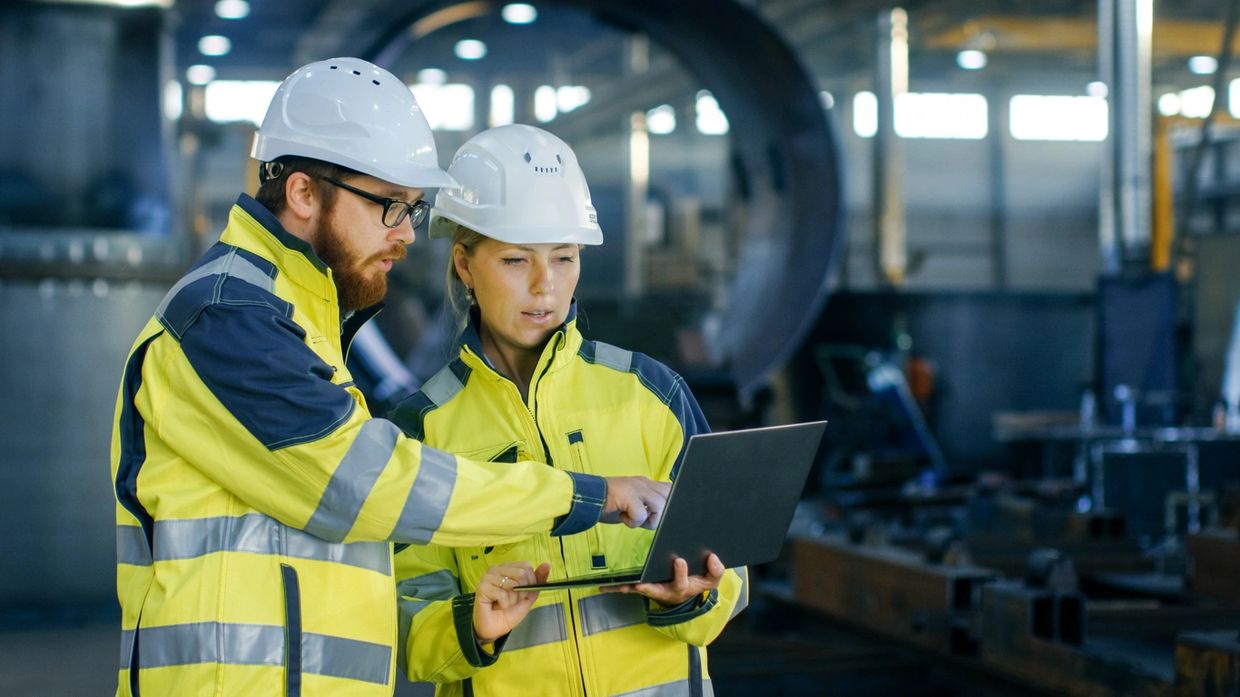 Two safety professionals in safety gear looking at schematics on laptop in factory setting