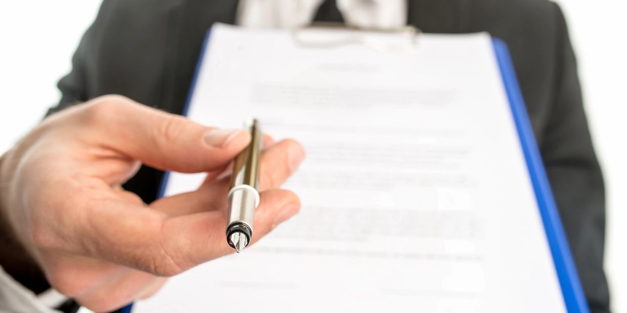 A real estate agent is handing someone a contract, with a pen, indicating for them to sign it. 