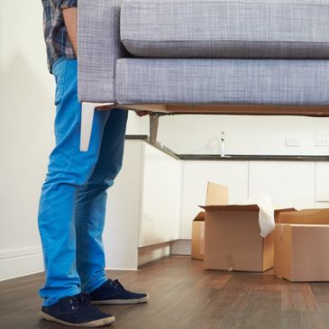 Person lifting a gray sofa in a room with moving boxes.