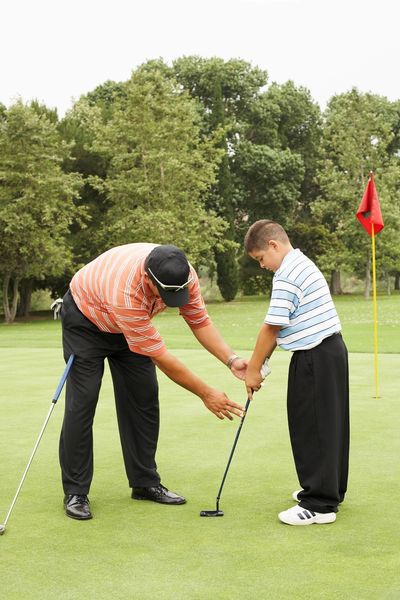 An adult teaching a boy how to putt on a golf green.