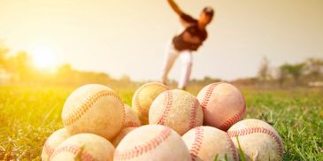 Pile of baseballs with a player pitching in the background at sunset.
